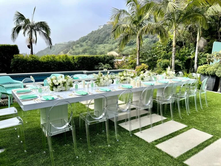 Long outdoor celebration table with white floral centerpieces and clear chairs