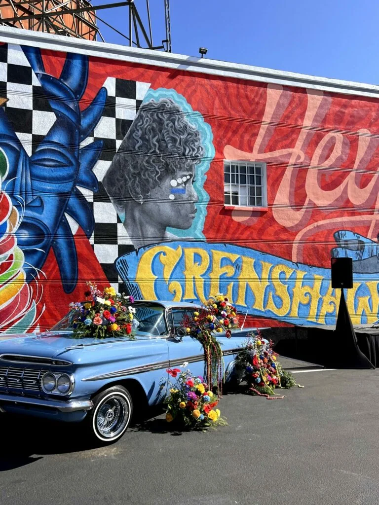 Wide view of a vintage blue car transformed with vivid floral installations beside a mural